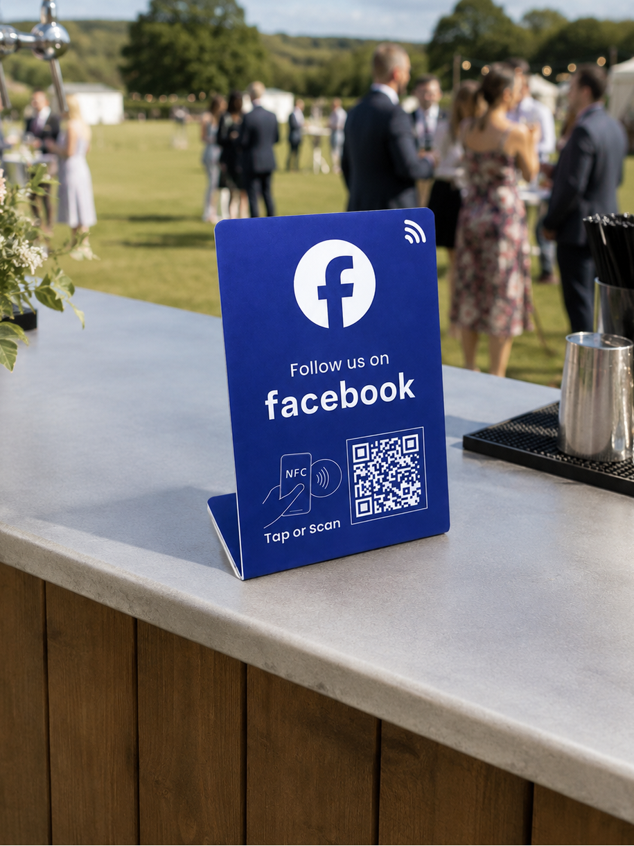 A blue freestanding sign on a bar counter at an outdoor event, with the Facebook logo and text reading 'Follow us on facebook' and a QR code. Guests are blurred in the background on a grassy field under a partly cloudy sky.